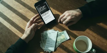 Overhead view of a person's hands holding a smartphone displaying news, with a gas receipt and bills on a worn wooden table, illustrating the impact of current events on personal finance.