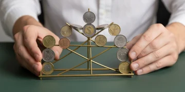 A person's hands carefully balancing various international coins on a brass rod structure, representing global economic interconnectedness on a dark green surface.