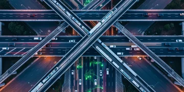 Aerial view of a complex urban transport network at dusk, with glowing green lines representing global trade routes intertwined with roads and railways, illustrating interconnectedness and economic shifts.