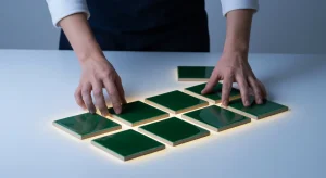 A person's hands arranging dark green ceramic tiles on a white surface, symbolizing proactive planning for a productive morning routine.