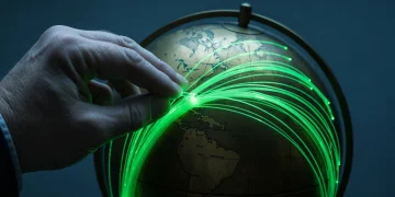 A weathered hand tracing glowing green fiber optic cables intricately crisscrossing an antique brass globe, symbolizing global economic connections and supply chains.