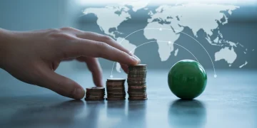 A human hand gently pushes a stack of copper coins across a concrete surface towards a green sphere, representing economic forces affecting personal finance.