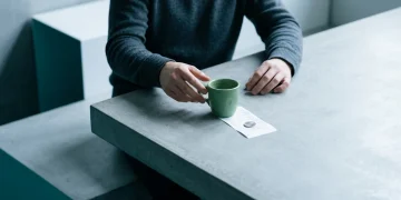 A person's hand hovering over a green coffee mug and a white receipt with a silver coin, illustrating cognitive biases in daily economic decisions.