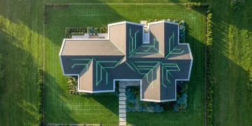 Aerial view of a futuristic modular smart home with glowing green energy patterns on its roof, surrounded by a lawn with embedded sensors, symbolizing autonomous living and predictive environments.